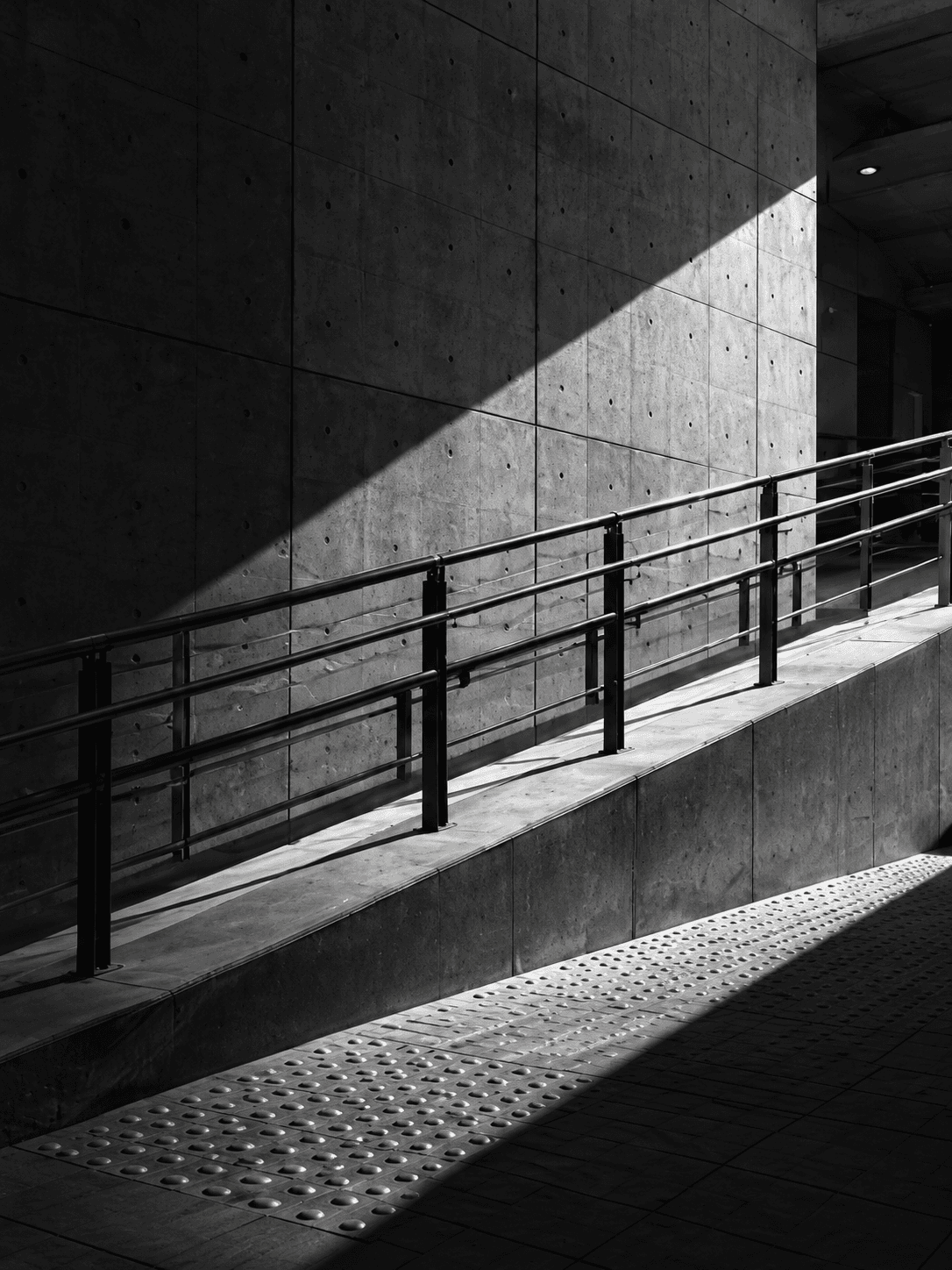 Une rampe longe un mur en béton, entre ombre et lumière.
